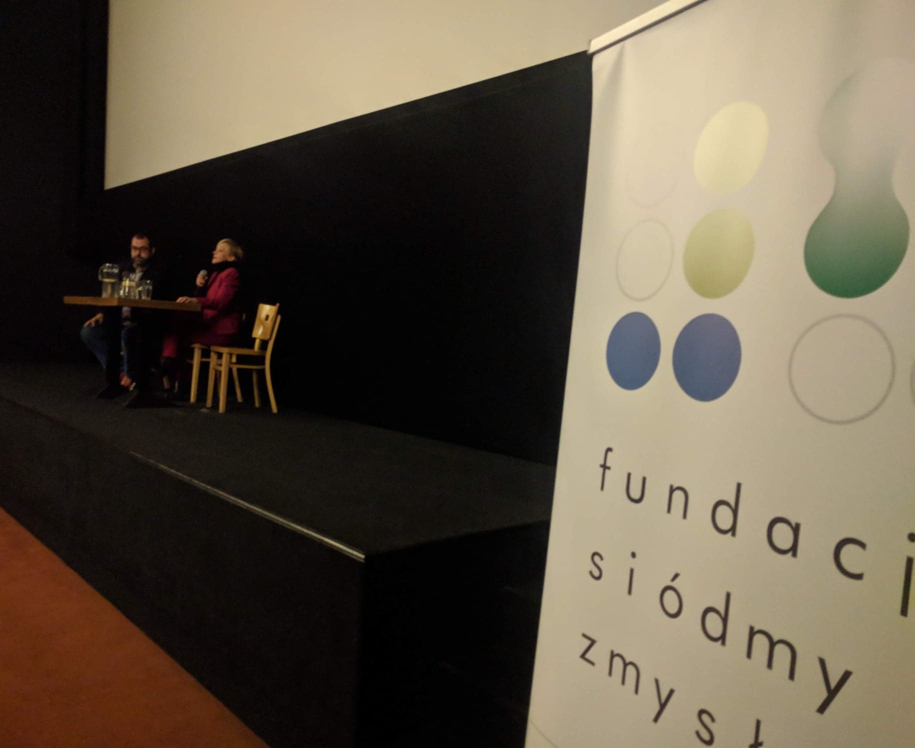 Scena pod białym kinowym ekranem. Przy stoliku siedzą Maciej Gil oraz Barbara Kurzaj. W lewym rogu baner Fundacji Siódmy Zmysł